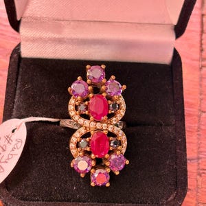 May include: A gold-toned ring with a cluster of gemstones, including oval rubies and round amethysts. The ring is set in a decorative design with small clear stones and is displayed in a black jewelry box.
