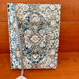May include: A rectangular journal with a fabric cover featuring a blue and white damask pattern. The cover is embellished with silver metallic thread embroidery and clear sequins arranged in a central floral motif. A grey ribbon bookmark is attached.