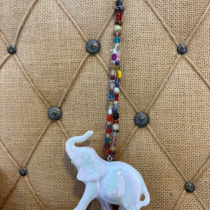 May include: A hanging ornament featuring a white iridescent elephant figurine suspended from a gold-toned metal hook by a long strand of multi-colored glass and metallic beads in various shapes, sizes, and hues including red, blue, and yellow.