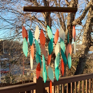 May include: A colorful glass wind chime with teal, white, and orange leaves hanging from a wooden dowel. The wind chime is hanging from a wooden deck railing.