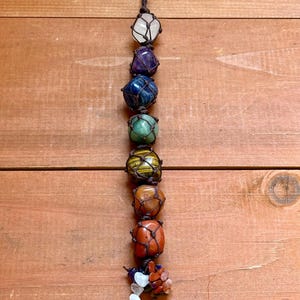 May include: A vertical arrangement of seven colorful gemstones, each encased in a knotted brown cord. The stones include clear quartz, amethyst, blue agate, green aventurine, tiger's eye, carnelian, and red jasper. The arrangement is suspended from a brown cord.