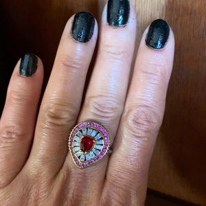 Op de afbeelding: Een gouden ring met een traanvormige rode steen omringd door kleinere roze en heldere stenen.
