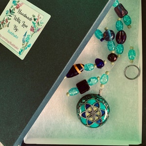 May include: A turquoise blue and gold geometric pendant necklace with a flower of life design. The necklace is in a black gift box with a sticker that says "Handmade with Love By: BuddhaArt etsy.com/shop/BuddhaArt".
