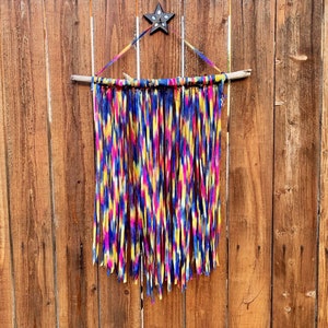 May include: A colorful wall hanging made of fabric strips tied to a branch. The fabric is a mix of blue, yellow, pink, and white with a tie-dye pattern.