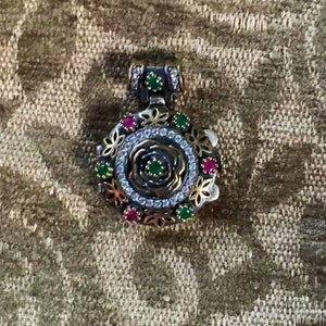 May include: A detailed pendant with a floral design. The pendant features a central rose motif, surrounded by a ring of small white stones, and accented with green and red gemstones. The metalwork includes gold and silver tones, with intricate floral patterns.