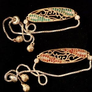 Puede incluir: Dos pulseras de oro con cadenas de plata y cierres ajustables. Las pulseras presentan un diseño ovalado con un trabajo de filigrana intrincado y una fila de piedras preciosas verdes y rojas.