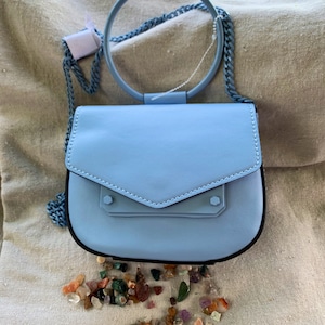 May include: A light blue satchel handbag with a circular top handle and a chain strap. The bag has a flap closure with a small pocket on the front. A scattering of colorful stones are in front of the bag.
