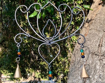 Op de afbeelding: Een metalen windgong met een decoratief ontwerp in de vorm van een hart en een traan. De gong heeft drie bellen en is versierd met gekleurde kralen.