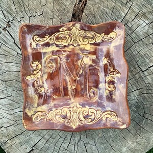 May include: A brown ceramic dish with a gold-coloured design. The dish has a square shape with rounded corners and features a large letter "M" in the centre. The dish is decorated with intricate floral patterns around the edges.