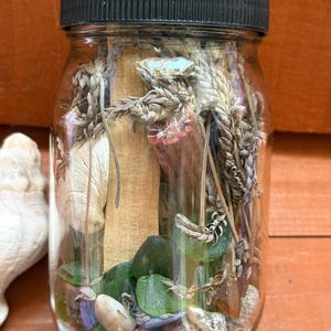 May include: A clear glass jar with a black lid and a red ribbon tied around the lid. The jar is filled with various items, including dried flowers, sea glass, and seashells.