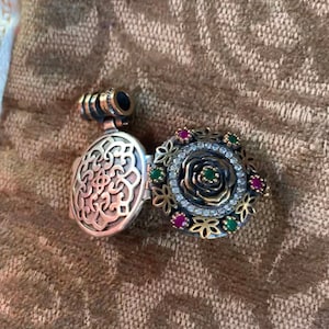 May include: A silver locket pendant with a filigree design on one side and a rose with green and red gemstones on the other side.
