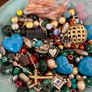 Op de afbeelding: Een kom gevuld met een verscheidenheid aan kleurrijke kralen, bedels en andere materialen voor het maken van sieraden. Er zijn hartvormige kralen, kruisbedels en een verscheidenheid aan andere vormen en maten. De kralen zijn gemaakt van glas, metaal en andere materialen.