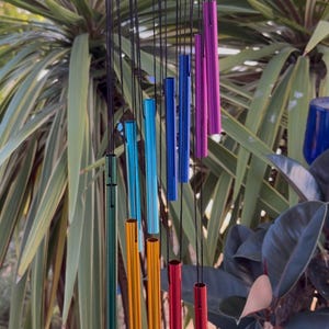 May include: A colorful wind chime with a dark red wooden top and a series of hanging metal tubes in various colors, including green, orange, red, blue, and purple. The tubes are suspended by black strings. The background features green foliage.