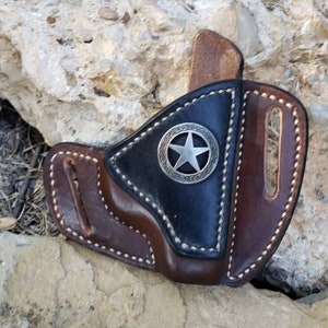 Op de afbeelding: Een bruine leren holster met een zwarte leren flap en een zilveren sterontwerp. De holster heeft witte stiksels rond de randen.