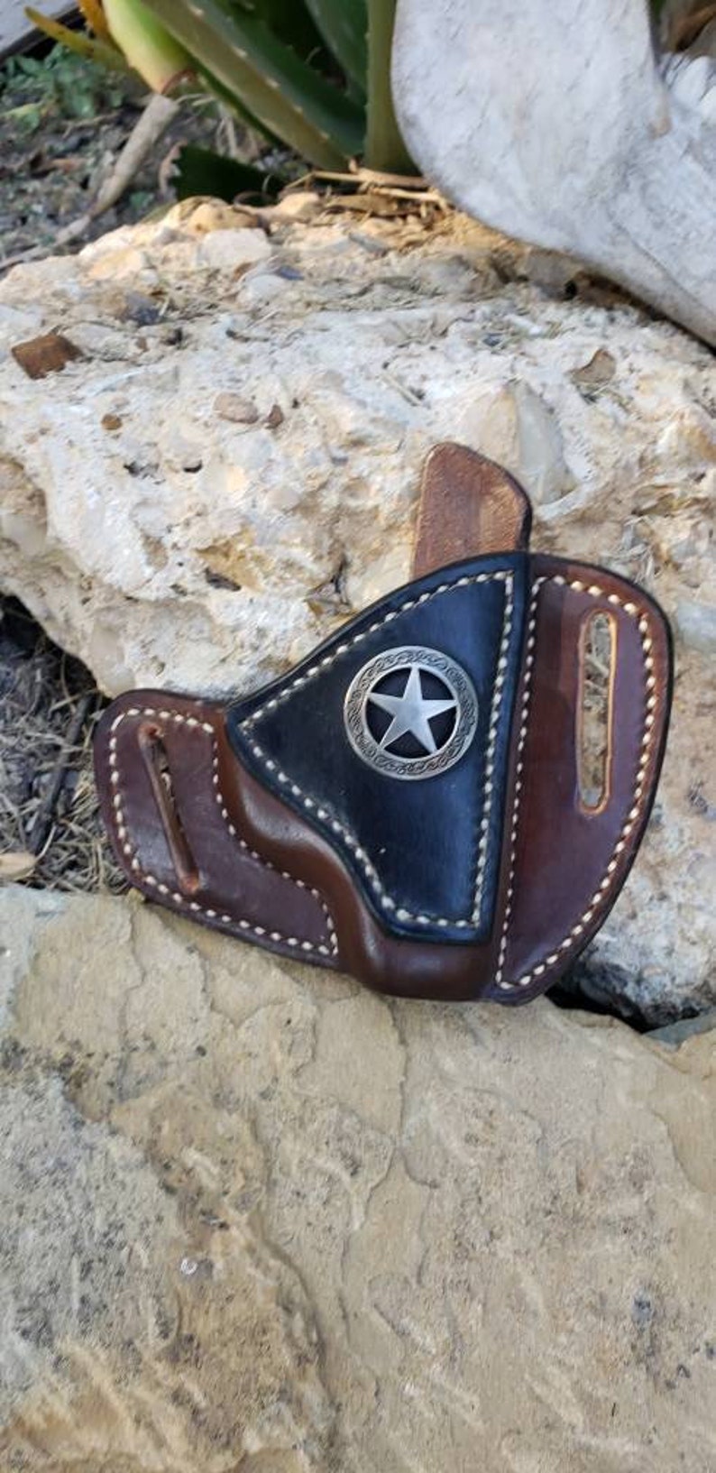 Op de afbeelding: Een bruine leren holster met een zwarte leren flap en een zilveren sterontwerp. De holster heeft witte stiksels rond de randen.