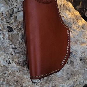 Puede incluir: Una funda de cuero marr&oacute;n con costuras blancas. La funda tiene un peque&ntilde;o logotipo en relieve que dice "Rugged Leather Goods".