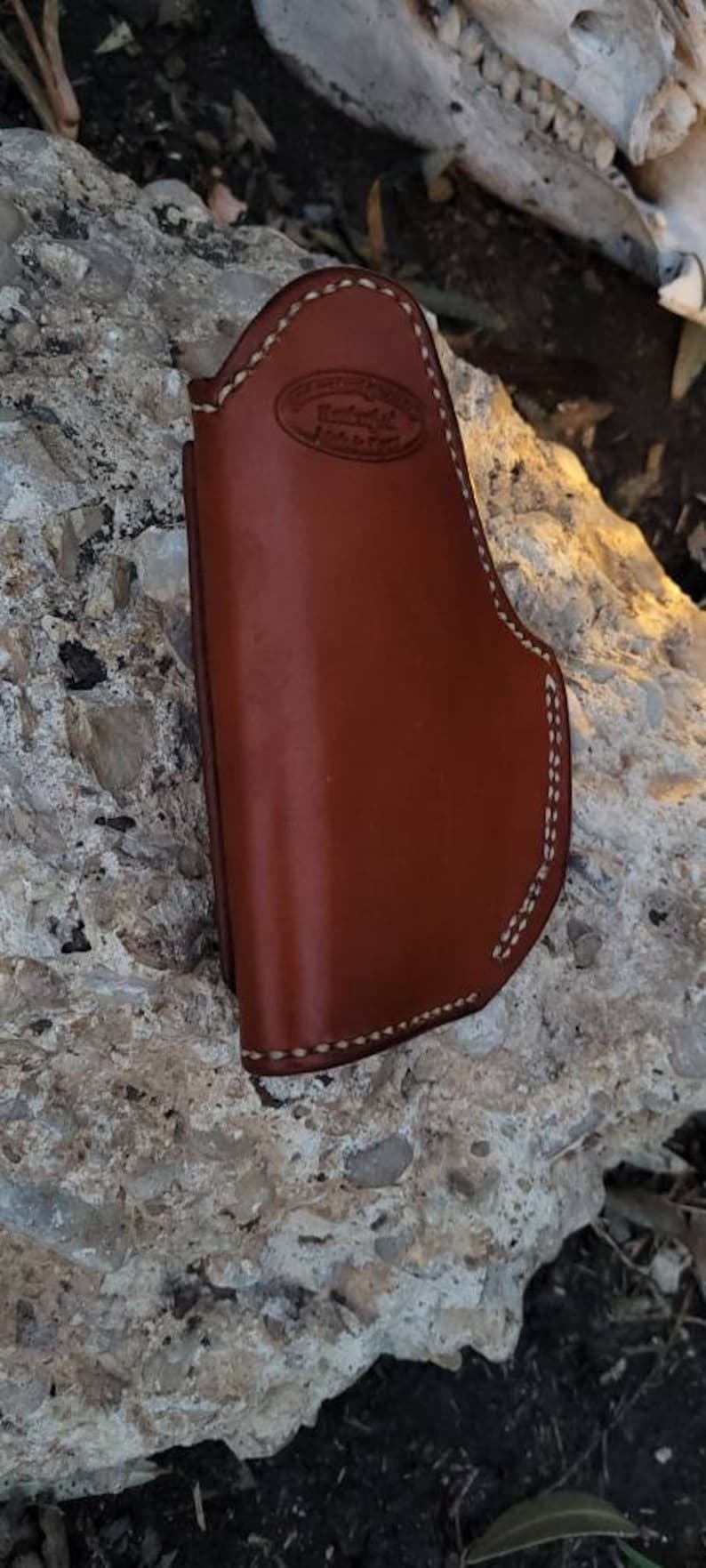Puede incluir: Una funda de cuero marr&oacute;n con costuras blancas. La funda tiene un peque&ntilde;o logotipo en relieve que dice "Rugged Leather Goods".