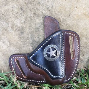 Op de afbeelding: Een bruine leren holster met een zwarte leren inzet en een zilveren sterontwerp. De holster heeft een gestikte rand en een riemlus.