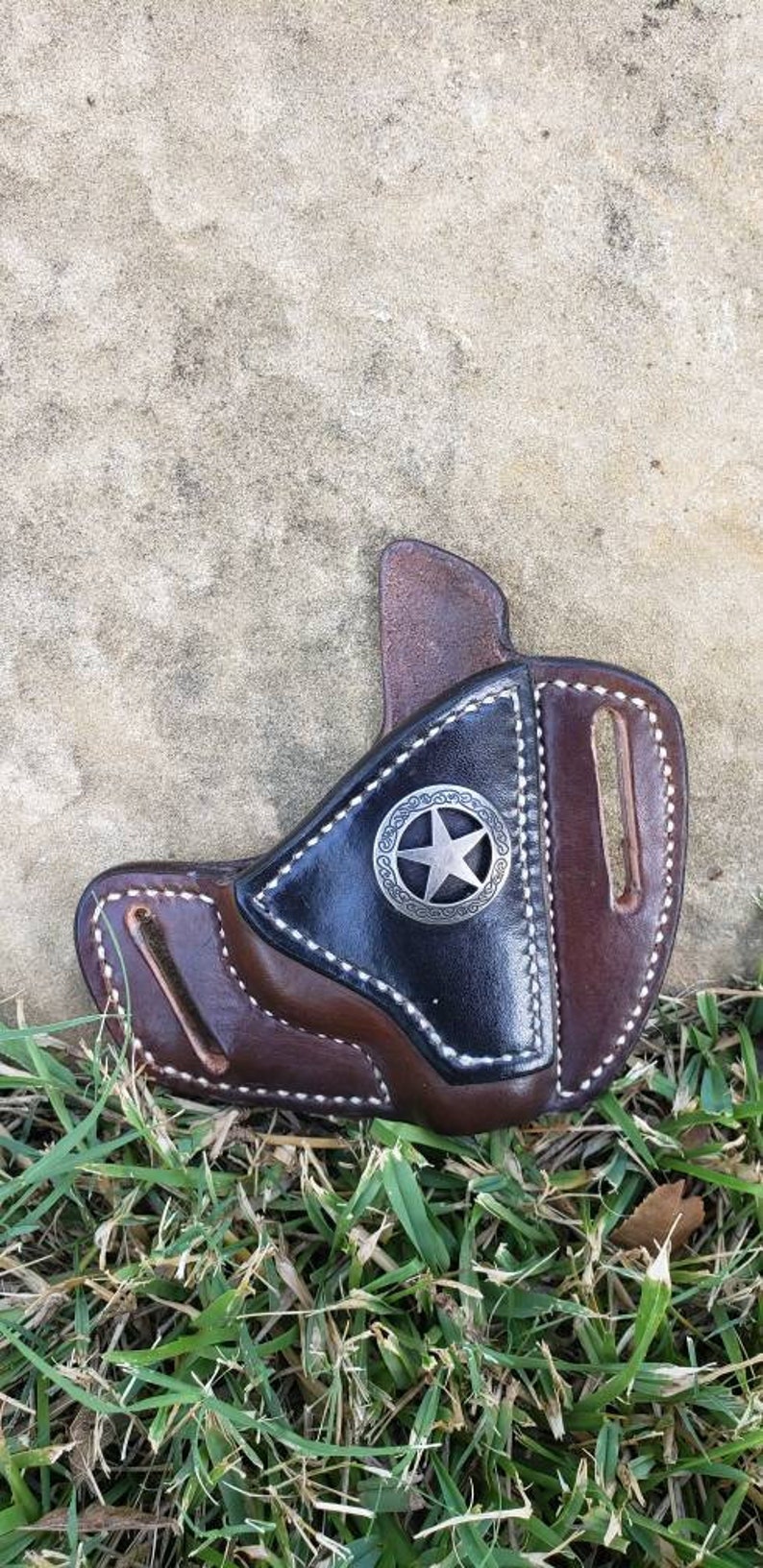 Op de afbeelding: Een bruine leren holster met een zwarte leren inzet en een zilveren sterontwerp. De holster heeft een gestikte rand en een riemlus.