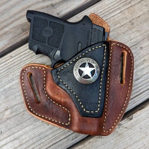 Op de afbeelding: Een bruine leren holster met een zwarte leren voering en een zilveren stervormig badge. De holster is ontworpen om een klein zwart pistool te houden. "Caution:  Loaded" is gedrukt op de zijkant van het pistool.