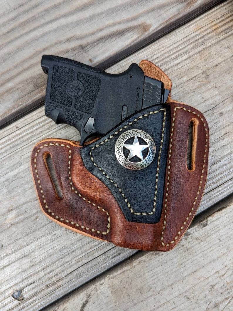 Op de afbeelding: Een bruine leren holster met een zwarte leren voering en een zilveren stervormig badge. De holster is ontworpen om een klein zwart pistool te houden. "Caution:  Loaded" is gedrukt op de zijkant van het pistool.