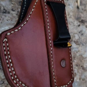 Puede incluir: Una funda de cuero marr&oacute;n con detalles de costura blanca y un clip de metal negro. La funda est&aacute; dise&ntilde;ada para sostener un arma de fuego.