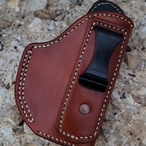 Puede incluir: Una funda de cuero marr&oacute;n con detalles de costura blanca y un clip de metal negro. La funda est&aacute; dise&ntilde;ada para sostener un arma de fuego.