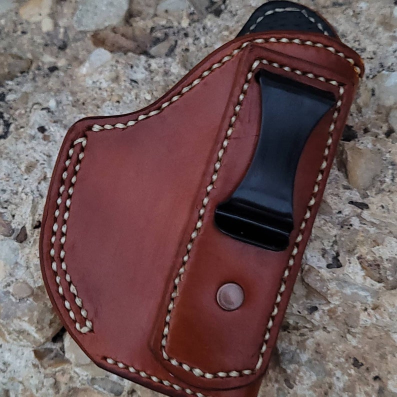 Puede incluir: Una funda de cuero marr&oacute;n con detalles de costura blanca y un clip de metal negro. La funda est&aacute; dise&ntilde;ada para sostener un arma de fuego.