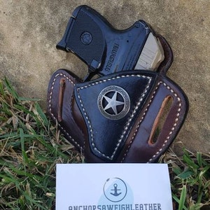 Op de afbeelding: Een bruine leren holster met een zwart sterontwerp en een zwart pistool erin. De holster is handgemaakt door AnchorSawWeighLeather, een bedrijf dat eigendom is van Jeffrey Altamirano. De visitekaartje ligt op een bed van groen gras.