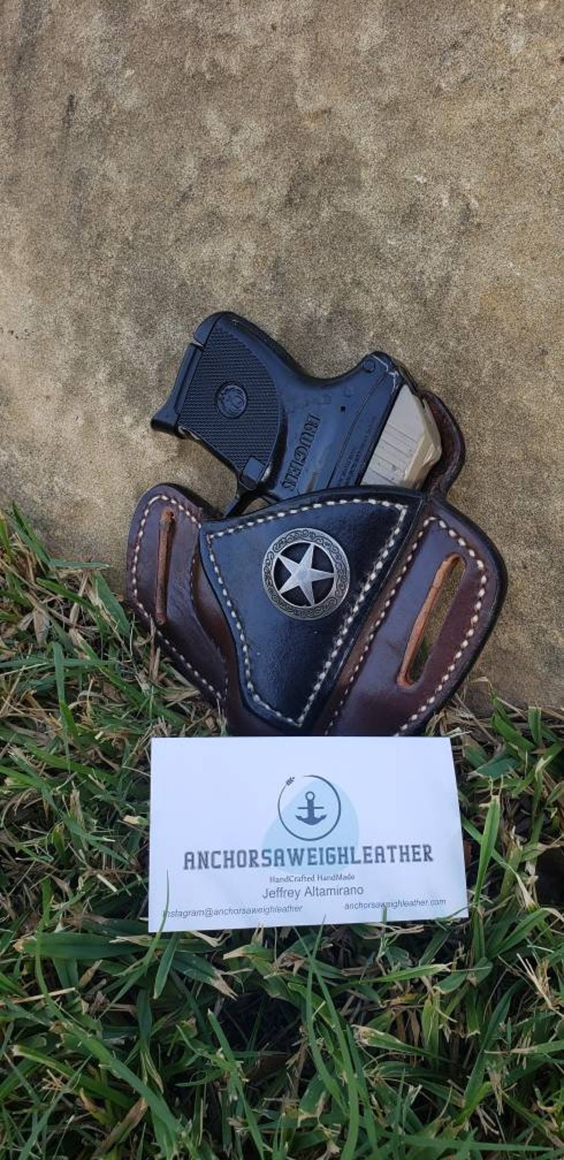 Op de afbeelding: Een bruine leren holster met een zwart sterontwerp en een zwart pistool erin. De holster is handgemaakt door AnchorSawWeighLeather, een bedrijf dat eigendom is van Jeffrey Altamirano. De visitekaartje ligt op een bed van groen gras.