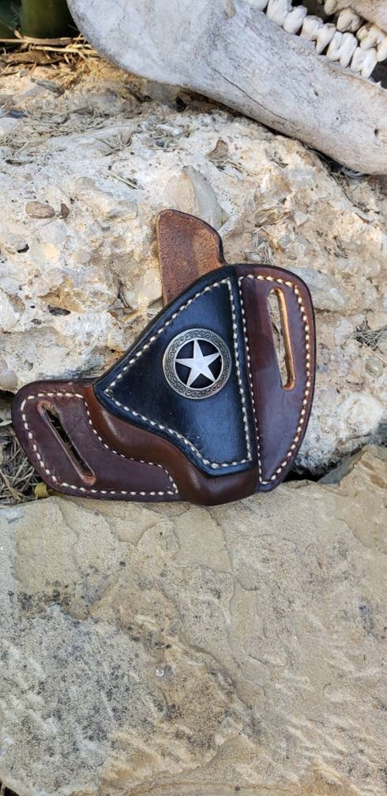 Op de afbeelding: Een bruine en zwarte leren holster met een zilveren sterontwerp. De holster is gemaakt van hoogwaardig leer en heeft een gestikt ontwerp.