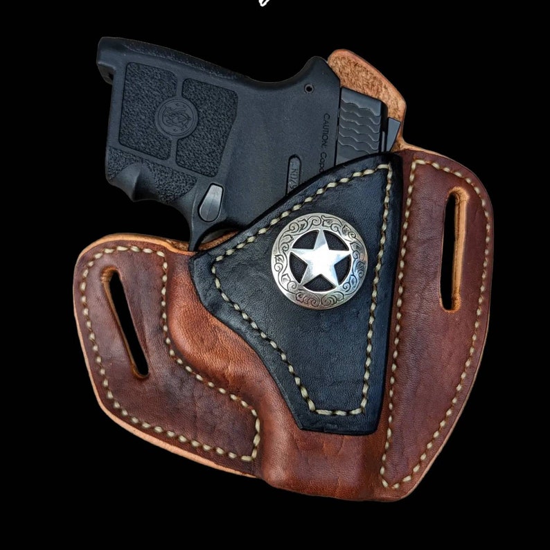 Op de afbeelding: Een bruine leren holster met een zwarte leren voering en een zilveren sterontwerp. Een zwart pistool zit in de holster.