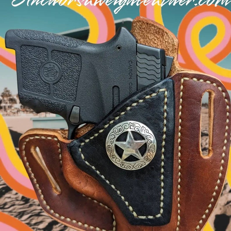 Op de afbeelding: Een bruine leren holster met een zwarte leren voering en een zilveren concho in de vorm van een ster. De holster is ontworpen om een klein zwart pistool te dragen. De tekst "CAUTION CAPAC" is zichtbaar op het pistool.