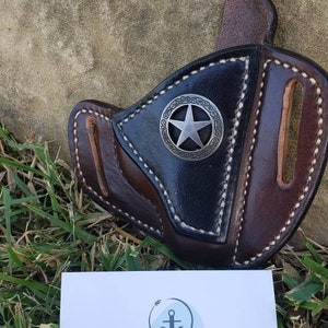 Op de afbeelding: Een bruine leren holster met een zwarte leren voering en een zilveren stervormig metalen accent. De holster is handgemaakt en gemaakt door AnchorSawWeighLeather. De visitekaartje bevat de tekst "Handcrafted Handmade Jeffrey Altamirano" en de Instagram-handle "@anchorsawweighleather".