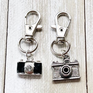 May include: Two silver camera keyrings with a silver swivel clasp. One camera charm is black with a rhinestone lens. The other camera charm is silver with a black lens.