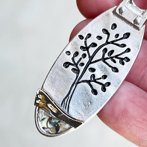 May include: A silver oval pendant featuring a black tree design and a gold and abalone shell accent.