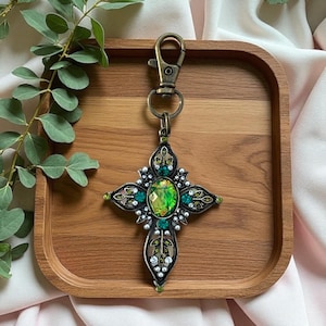 May include: A decorative cross-shaped keychain with a bronze-colored metal finish. The cross features a large, oval-shaped green gemstone at its center, surrounded by smaller green and clear gemstones, and faux pearls. The keychain is on a wooden tray.