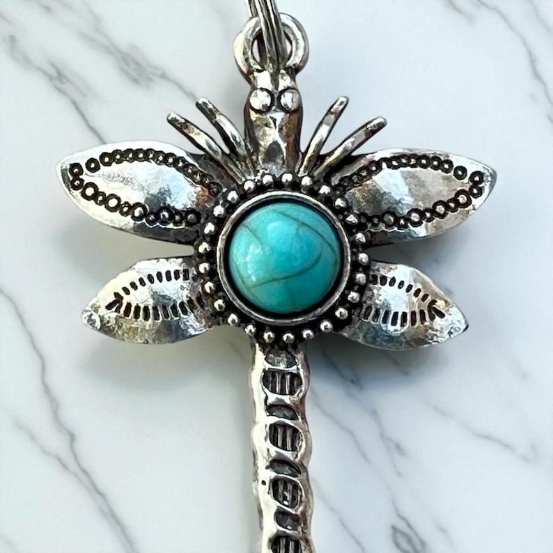 May include: Silver-toned dragonfly pendant with a turquoise-colored stone center. The dragonfly's wings are detailed with small, circular patterns. The pendant is suspended from a silver chain.