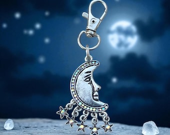 May include: Silver-toned keychain featuring a crescent moon with a face and dangling stars. The moon is adorned with iridescent crystals. The keychain has a clasp for easy attachment. The background shows a night sky with a full moon.