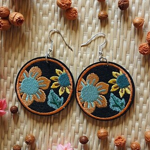 May include: A pair of round black earrings with embroidered floral designs. Each earring features a teal and orange flower with yellow accents, framed by an orange border. The earrings hang from silver-colored hooks.