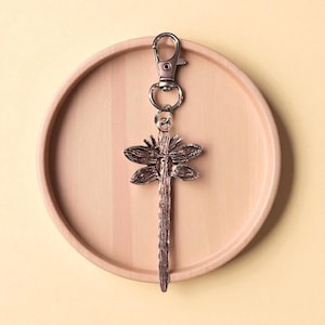 May include: A dragonfly-shaped keychain with a silver clasp and ring. The dragonfly pendant is bronze-colored and detailed, resting on a light brown wooden tray. The background is a solid, pale yellow.