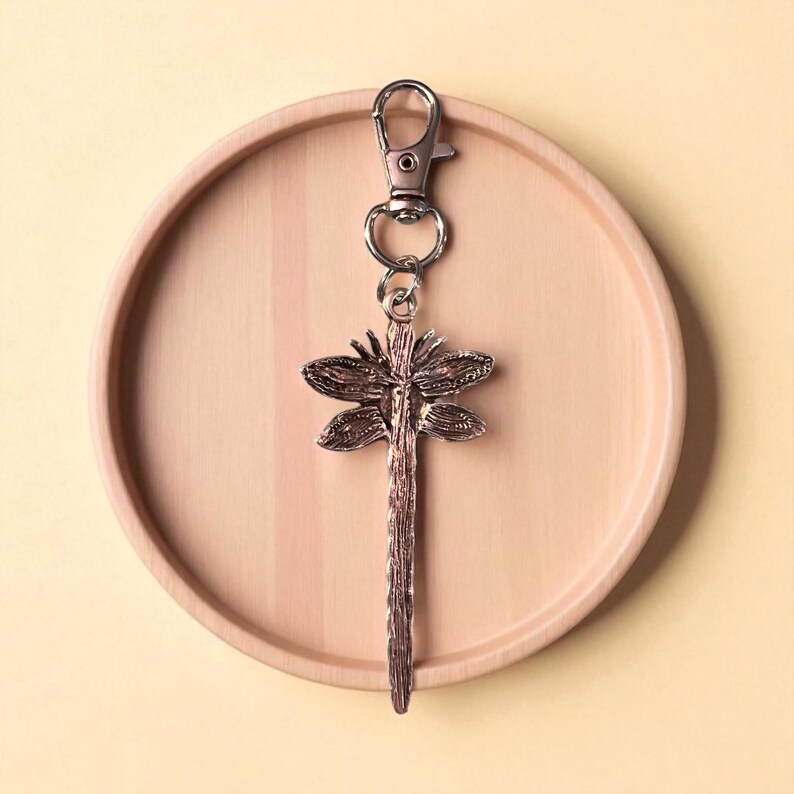 May include: A dragonfly-shaped keychain with a silver clasp and ring. The dragonfly pendant is bronze-colored and detailed, resting on a light brown wooden tray. The background is a solid, pale yellow.
