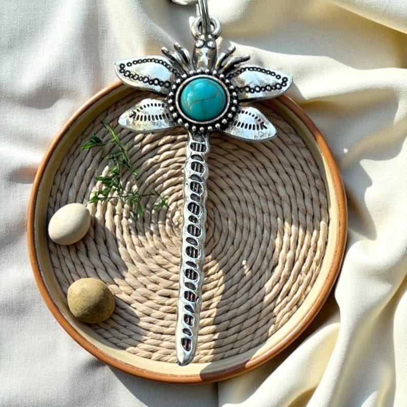 May include: A silver dragonfly pendant with a turquoise center stone, displayed on a woven tray with two small stones and greenery. The dragonfly has detailed wings and a textured body, creating a bohemian aesthetic.
