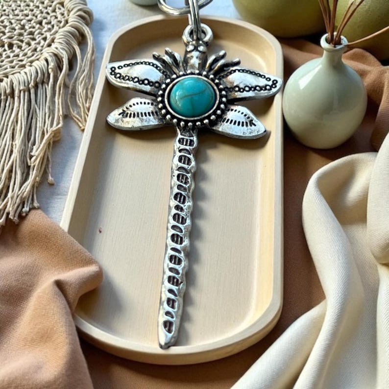 May include: A silver-colored dragonfly pendant with a turquoise-colored stone in the center. The dragonfly has detailed wings and a long, textured body. The pendant is on a light-colored tray, with a decorative macrame piece and a small vase in the background.