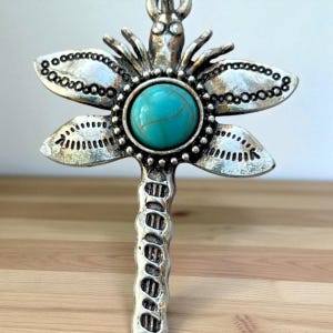 May include: A silver-toned dragonfly pendant with a turquoise-colored stone center. The dragonfly's wings are detailed with small, circular accents. The pendant has a textured, antique finish, and is likely a piece of jewelry.