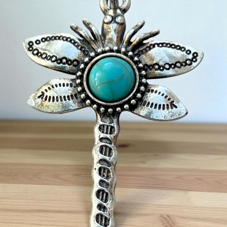 May include: A silver-toned dragonfly pendant with a turquoise-colored stone center. The dragonfly's wings are detailed with small, circular accents. The pendant has a textured, antique finish, and is likely a piece of jewelry.