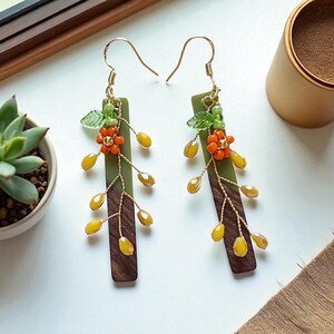 May include: A pair of dangle earrings featuring a botanical design. Each earring has a dark brown wooden rectangle base, topped with a green rectangle. Delicate gold wire branches hold orange flower clusters and yellow teardrop-shaped beads. The earrings have gold-colored ear wires.