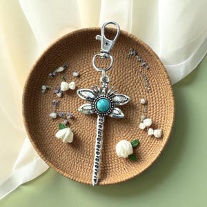 May include: Silver-toned dragonfly keychain with a turquoise center stone. The dragonfly has detailed wings and a long, textured body. The keychain is attached to a silver clasp. The item is displayed on a woven, circular tray with floral accents.