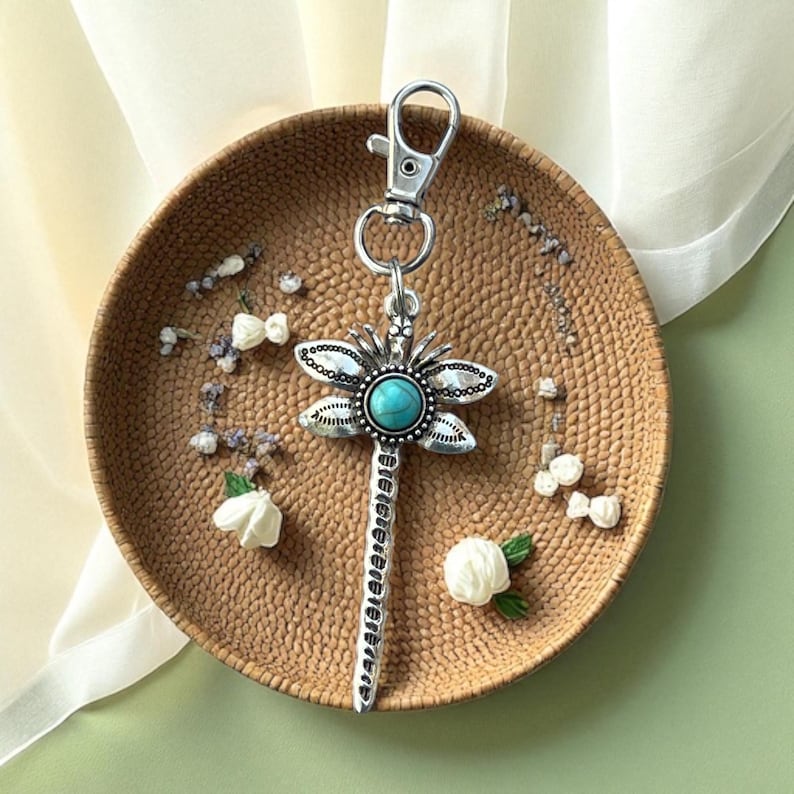May include: Silver-toned dragonfly keychain with a turquoise center stone. The dragonfly has detailed wings and a long, textured body. The keychain is attached to a silver clasp. The item is displayed on a woven, circular tray with floral accents.