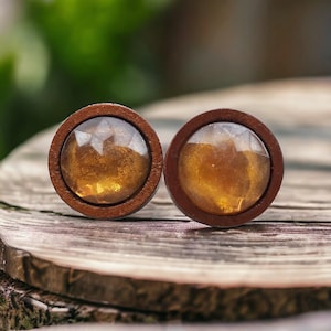 May include: A pair of circular stud earrings featuring a dark brown wooden frame surrounding a faceted, translucent amber-colored resin center with a shimmering, golden-brown metallic flecked interior finish.